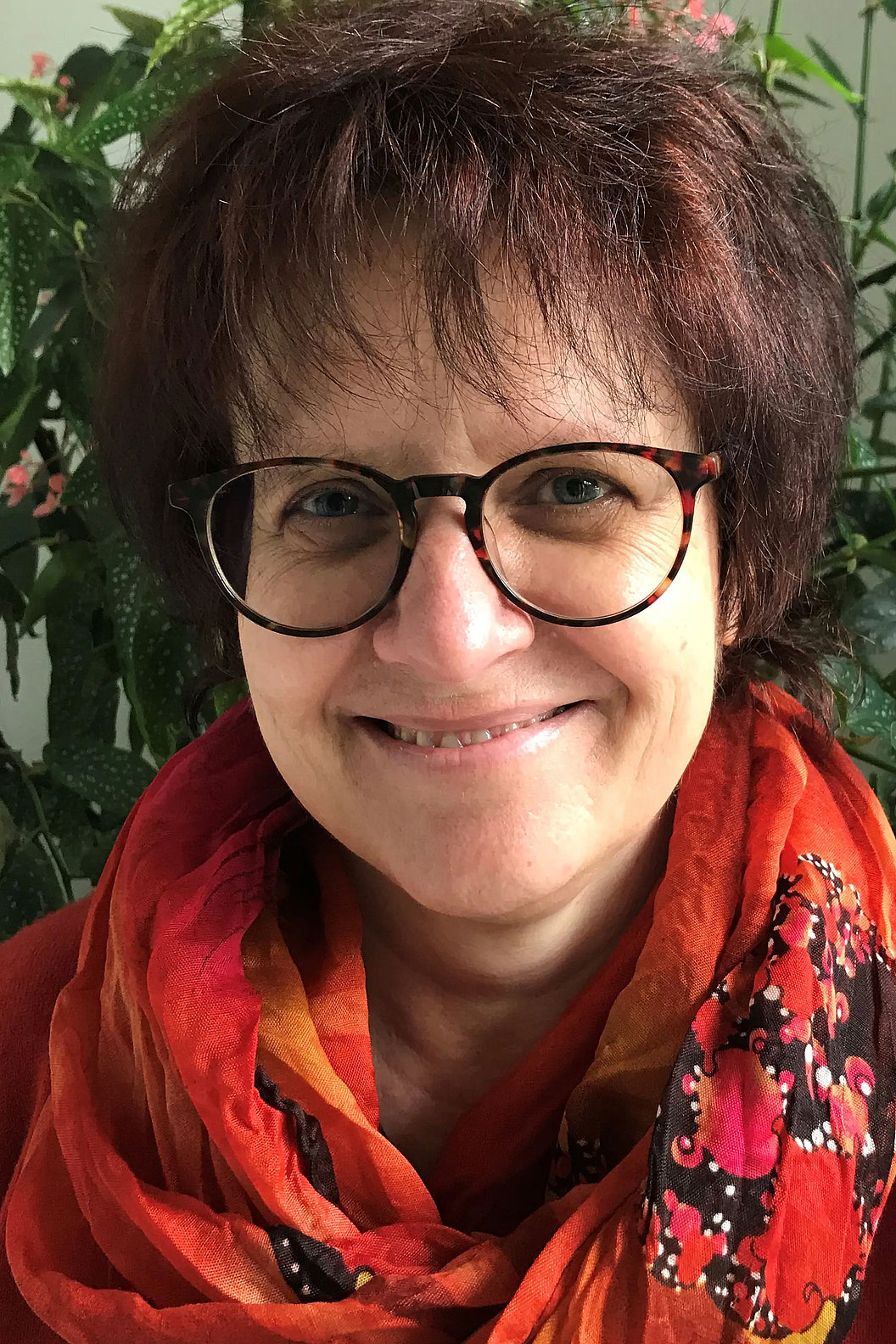 Dr. Karin Rodrian mit kurzen braunen Haaren trägt eine orange-rote Schal mit schwarzem und rotem Muster vor grünem Pflanzenhintergrund.