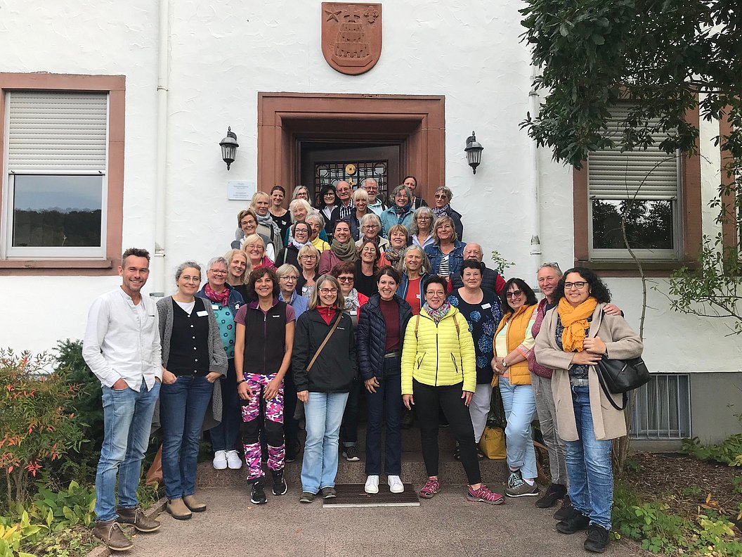 Gruppe von Menschen vor einem Gebäude mit Wappen über dem Eingang, umgeben von Fenstern und Pflanzen.