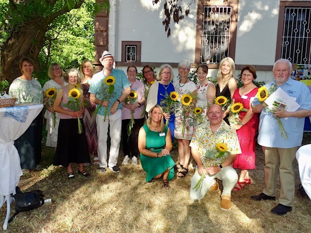 Gruppe von Menschen vor einem Haus, die jeweils eine Sonnenblume halten, zwei Personen knien vorne auf dem Gras